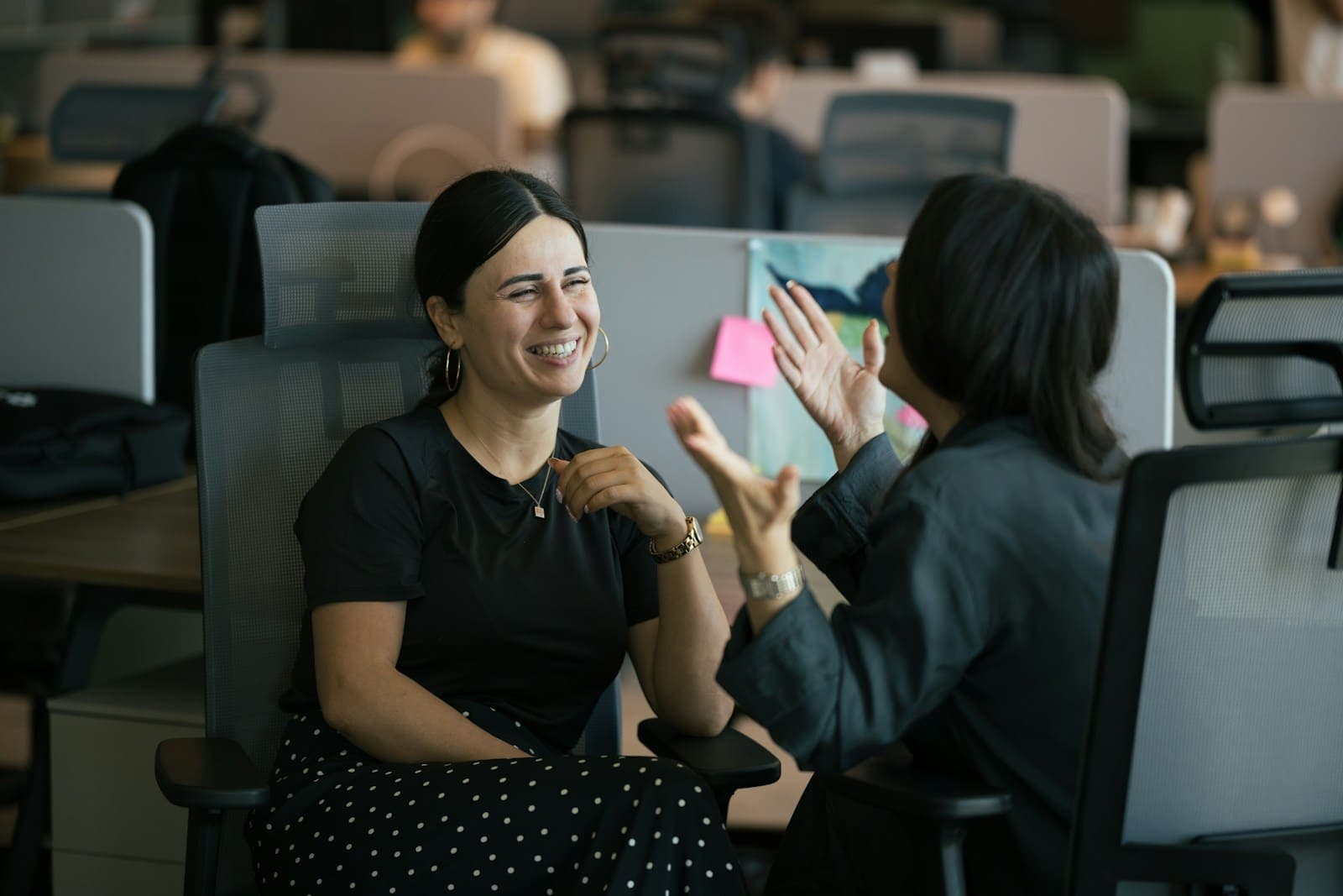 Two colleagues laughing as they chat across a desk in an open-plan office
